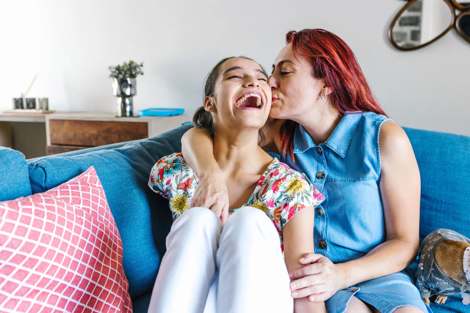 A mother kissing her daughter on the cheek as she laughs, sitting together on the couch at home