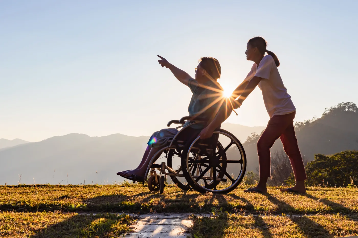 A man in a wheelchair and a support worker outdoors at sunset, looking out across the hills